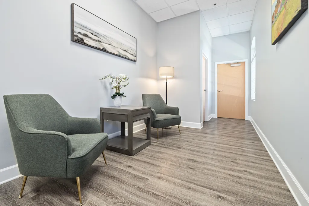The image displays the Turning Point of Tampa Drug and Alcohol Detox Waiting Area, a modern and simplistic waiting area or reception room. The room features light gray walls, complemented by dark gray baseboards and a durable, wood-look laminate flooring in a gray-brown tone. There are two stylish, mid-century modern inspired armchairs with a textured teal upholstery and angled gold legs. Between the chairs is a contemporary dark wooden side table that supports a white vase with white orchid flowers. Above the side table, on the wall, is a panoramic abstract painting with earthy tones and streaks of white, which could be interpreted as a landscape. A floor lamp with a classic shade design stands in the corner, emitting a warm light. To the right, another abstract painting with more vibrant colors hangs on the wall, near an exit door with a wooden finish. The overall atmosphere is clean, professional, and inviting, likely designed to put visitors at ease while they wait.