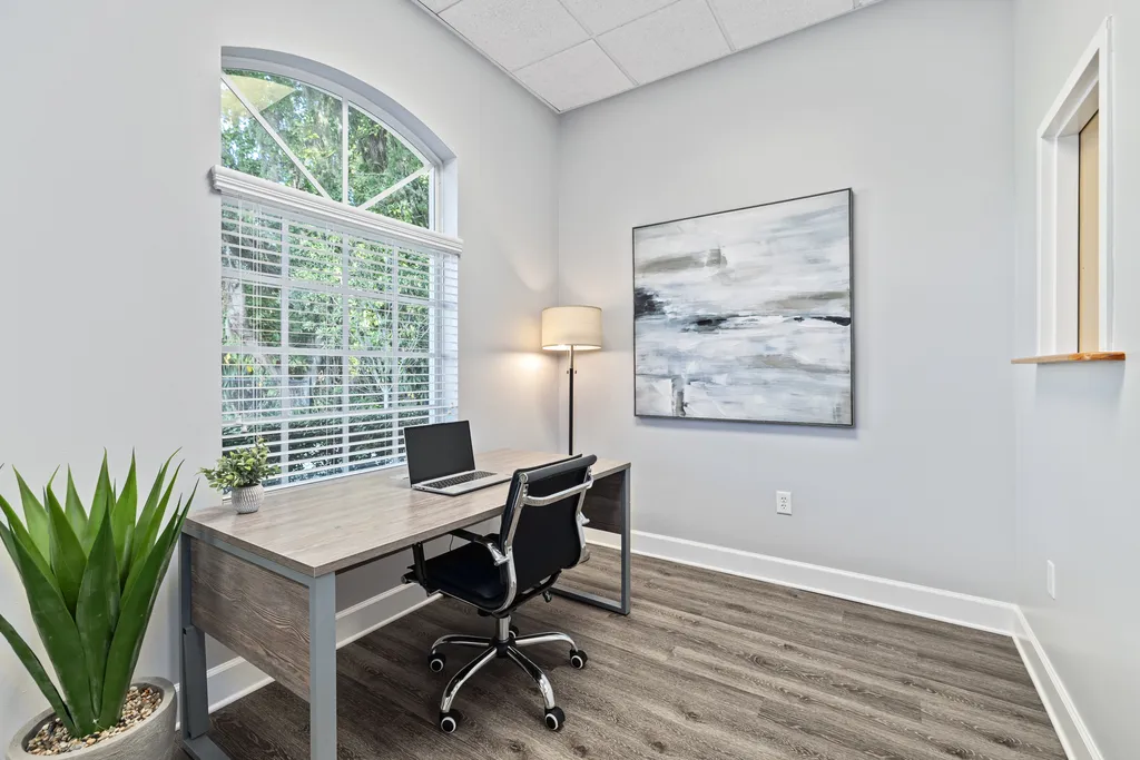 Turning Point of Tampa Detox desk for clinicians. A space that is designed to be both functional and aesthetically pleasing. A large arched window with white blinds fills one wall, providing a view of greenery outside and allowing natural light to brighten the room. In the corner, there’s a sleek desk with a wood-like finish, accompanied by a modern office chair on wheels, indicating a workspace. A laptop open on the desk suggests that the space is in use. To the left of the desk, a large potted plant adds a lively touch of green, complementing the view outside and enhancing the room's natural ambiance. On the wall, a large abstract painting in grayscale tones hangs, providing a sophisticated visual interest that ties in with the room's modern decor. A floor lamp stands to the left, offering additional lighting options. The room's color scheme is neutral, with light gray walls and wood-look flooring that continues the modern, clean aesthetic seen throughout the other images of the location. A small, decorative shelf is mounted on the right wall, holding a small potted plant, which adds a personal touch to the workspace. The overall impression is of a tranquil and contemporary office setting, designed for focus and productivity, while still offering elements of nature and art to create a pleasant working environment.