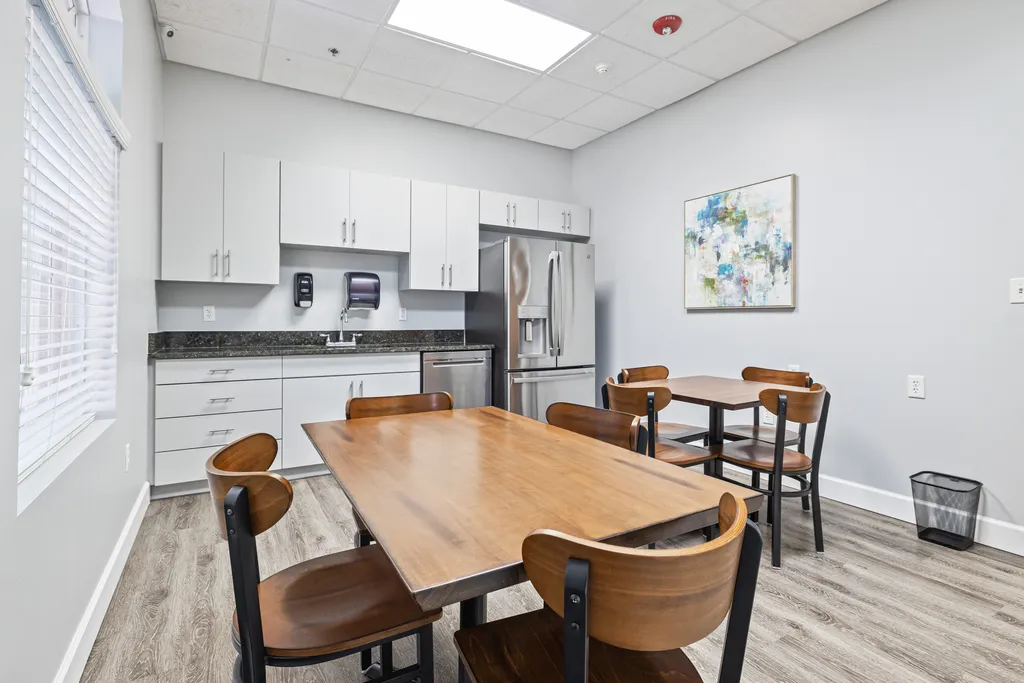Turning Point of Tampa Detox Kitchen. This image showcases a different angle of what appears to be an office break room or kitchenette area. The focus is on a large wooden dining table surrounded by four chairs with a dark wood finish and black metal supports. This table is likely used for eating, informal meetings, or as a communal workspace. The kitchen area features modern, white upper and lower cabinets, and drawers with sleek, silver handles that provide ample storage. The countertops are a dark granite or marble, and house various appliances such as a coffee maker, a paper towel dispenser, and a stainless steel double-door refrigerator, indicating that this is a fully functional kitchenette. There is a large abstract painting on the wall, adding a splash of color and artistic flair to the room. The room is well-lit by natural light from the window on the left and overhead fluorescent lights. Like the previous images, the room has wood-like vinyl flooring, which is practical and easy to maintain. A trash bin is visible to the right, and the general cleanliness and orderly setup suggest that the space is well-maintained. The ceiling has a fire safety feature (likely a smoke detector), and the overall atmosphere is clean, bright, and modern, designed for the comfort and utility of the people using the space.
