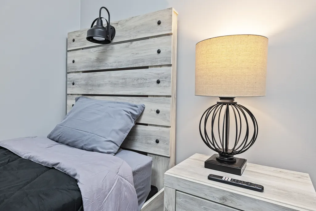 Turning Point of Tampa Detox Bedroom. This close-up image provides a detailed look at one of the beds and the accompanying nightstand in the bedroom setting previously described. The weathered wooden headboard is prominently displayed, with a wall-mounted black reading light positioned just above it, adding to the functional yet stylish décor. The nightstand is also made of the same weathered wood, and it features two drawers with black handles that match the reading light. On top of the nightstand is a decorative lamp with a black, cage-like base and a square pedestal, holding a beige lampshade that casts a warm glow. This lamp design adds an element of sophistication to the room. Beside the lamp is a remote control, suggesting that the room includes modern amenities like a television. The linens on the bed are consistent with the previous images, showcasing a neat arrangement with a dark grey comforter and light grey sheets. The overall aesthetic is contemporary with a touch of industrial design, aimed at providing a comfortable and inviting atmosphere for rest.