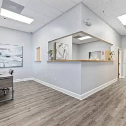 Turning Point of Tampa Detox Nurses Station. This image shows a bright and spacious interior, characterized by its clean and professional design. On the left, there is a desk with a modern design, a desk lamp, and a large, healthy potted plant, which adds a touch of greenery to the room. Above the desk hangs a large abstract painting with streaks of gray and white, providing a contemporary artistic touch. In the middle of the room, there’s a reception counter with a natural wood trim, which creates a welcoming focal point. Above the counter is a large open window, allowing for visibility and communication between the front desk area and the adjoining space. To the right of the reception counter is a hallway with several doors, indicating multiple rooms or offices. The hallway is well-lit, likely by a combination of natural light from the skylight above and artificial lighting, ensuring a bright atmosphere conducive to work. The flooring throughout the space is a wood-like laminate or vinyl, consistent in pattern, suggesting easy maintenance and durability. The walls are painted a soft light color, which, along with the ceiling lights, enhances the open and airy feeling of the interior. Overall, the design of the room is geared towards functionality and efficiency, typical of a professional environment where clients or patients are received and attended to. The decor is minimal yet thoughtful, with artwork and plants to create a welcoming space while maintaining a professional ambiance.